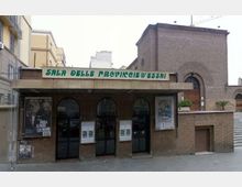 Facciata di un edificio in mattoni con ingresso coperto da una pensilina e l’insegna “Sala delle Province d’Essai”. Sotto la tettoia si vedono tre porte ad arco in vetro e bacheche con locandine, con un volume laterale più alto in mattoni e una piccola scala sul lato destro.