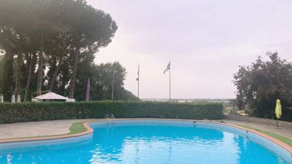 Piscina all’aperto dalla forma curva con acqua azzurra e bordo in pietra, circondata da siepi e alberi alti. Sullo sfondo si vedono un gazebo, aste con bandiere e un’area paesaggistica aperta sotto un cielo nuvoloso.