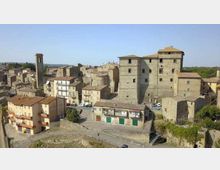 Veduta panoramica di un borgo in pietra con case addossate e una grande struttura fortificata simile a un castello al centro, affiancata da una torre campanaria. In primo piano si vedono una strada e un terrapieno, con colline verdi sullo sfondo sotto un cielo sereno.