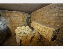 Interno sotterraneo in pietra con soffitto basso e pareti in blocchi, illuminato debolmente. Sulla destra c’è un sarcofago in pietra con rilievi decorativi e al centro un basamento con frammenti di pietra.