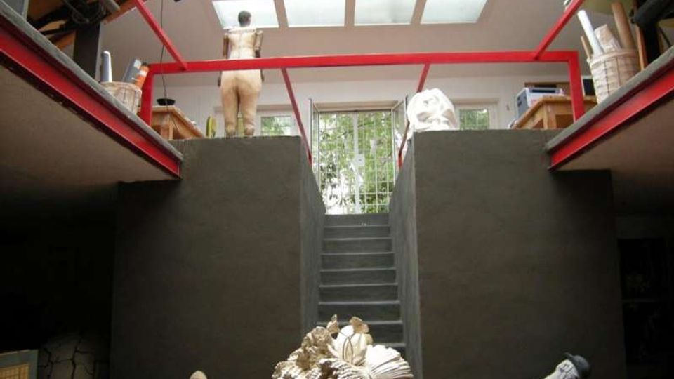 Interno di uno studio d’arte su due livelli con travi rosse a vista, grande lucernario a griglia sul soffitto e scala centrale tra pareti scure. Ai lati ci sono soppalchi con scaffali e materiali, e sul piano inferiore sono visibili sculture e oggetti artistici.