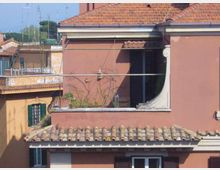 Facciata di un edificio residenziale con pareti rosa e tetto in tegole di terracotta, con una terrazza al primo piano delimitata da parapetto e ringhiera metallica. Sullo sfondo si vedono altri tetti e antenne in un contesto urbano.