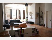 Soggiorno open space in stile loft con pavimento in legno, grandi finestre con tende scure e arredi moderni (divano, tavolino e pouf). Sulla destra una scala a chiocciola in metallo conduce a un soppalco, con pareti bianche decorate da una composizione di quadri.