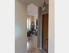 Vista da un corridoio verso un salotto con pavimento in piastrelle decorative a motivi geometrici. Si vedono una grande finestra, una libreria bianca, sedie imbottite, piante in vaso e una lanterna a sospensione, con una porta in legno sulla destra.