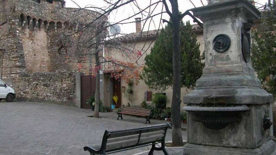 Piccola piazza lastricata con panchine e alberi spogli, affiancata da un edificio in pietra con muratura antica e aperture ad arco. In primo piano si vede una fontana monumentale in pietra con basamento squadrato e decori circolari.