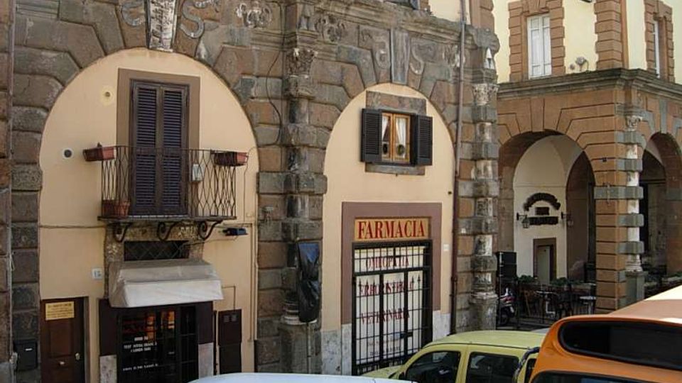Facciata di un edificio storico con grandi archi in pietra e pareti intonacate, con finestre a persiane e un piccolo balcone in ferro battuto. Al piano strada si vede l’ingresso di una farmacia con insegna “FARMACIA” sotto un portico ad archi; in primo piano sono parcheggiati alcuni veicoli.