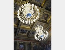Soffitto di una sala storica riccamente decorato con riquadri dipinti e cornici dorate, con grandi lampadari in vetro bianco sospesi. Sulle pareti sottostanti si intravedono affreschi e archi in stile classico.