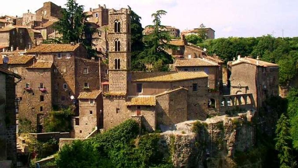 Borgo storico in pietra arroccato su uno sperone roccioso, con case addossate dai tetti in coppi e una torre campanaria centrale con finestre ad arco. Sullo sfondo si vede una collina boscosa e un ponte ad arcate che collega alcuni edifici sul margine del paese.