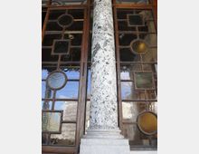 Dettaglio di una facciata con grande finestra a riquadri in legno e inserti di vetro colorato circolari e quadrati. Al centro si vede una colonna in marmo screziato su base chiara, davanti al serramento.