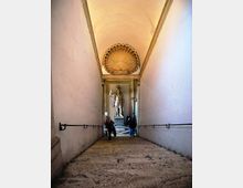 Scalinata in pietra all’interno di un edificio storico, con pareti chiare e corrimano in metallo che conduce a un vestibolo. In fondo si vede una grande statua su piedistallo e un soffitto decorato con una rosetta a conchiglia.