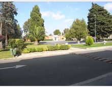 Strada asfaltata con aiuole curate e siepi basse, circondata da alberi alti e palme in un’area verde. Sullo sfondo si vedono edifici bassi e un’area di parcheggio con lampione e piccole fontane ornamentali.