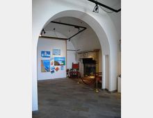 Interno di una sala espositiva con soffitto a volte e grande arco bianco, pavimento in pietra e illuminazione a binari. Sulla sinistra c’è una parete con disegni e dipinti appesi, mentre sulla destra si vede un grande camino in pietra delimitato da cordoni rossi.