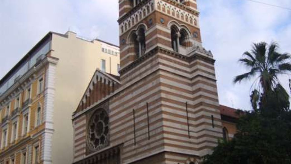 Chiesa in stile romanico-gotico con facciata a fasce orizzontali bianche e rosse e alto campanile con logge ad archi sovrapposti. L’edificio si affaccia su una piazza urbana, con palazzi adiacenti e una palma sul lato destro.