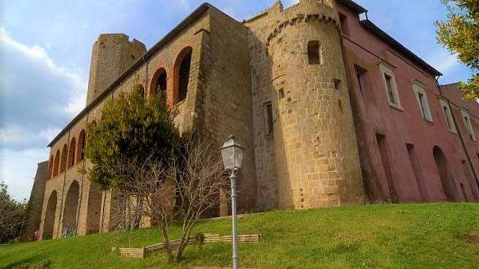 Edificio storico in pietra simile a un castello, con torre cilindrica e una lunga facciata ad archi, affiancato da una sezione intonacata rosa con finestre. In primo piano c’è un pendio erboso con alberi e un lampione, sotto un cielo parzialmente nuvoloso.