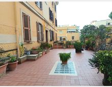 Ampia terrazza condominiale con pavimentazione in mattoni rossi, fioriere e vasi di piante lungo i lati e una griglia di vetro al centro. A sinistra si vede la facciata di un edificio con finestre e persiane, mentre sullo sfondo compaiono altri palazzi e alberi.