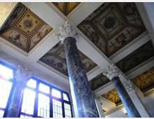 Interno di un edificio storico con alte colonne in marmo scuro e capitelli decorati, affacciate su grandi finestre che lasciano entrare molta luce. Il soffitto a cassettoni è suddiviso da travi bianche e presenta riquadri affrescati con motivi ornamentali.