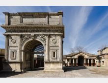 Arco monumentale in pietra con grande apertura a volta cassettonata, colonne laterali e bassorilievi decorativi sulla facciata. Davanti si apre un’ampia piazza lastricata, con sullo sfondo un portico con colonne e tetto in coppi sotto un cielo azzurro.