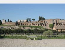 Veduta di un ampio complesso archeologico in muratura con resti di edifici e arcate su un pendio, con cipressi e altre alberature sullo sfondo. In primo piano si vede un’area verde delimitata da una staccionata e un sentiero ghiaioso con una panchina.