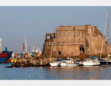 Rovina di una grande costruzione in pietra su un molo, con pareti parzialmente crollate e piccole aperture, affacciata su un porto. In primo piano si vede l’acqua con barche a vela e piccole imbarcazioni ormeggiate, mentre sullo sfondo sono presenti altre navi e strutture portuali.