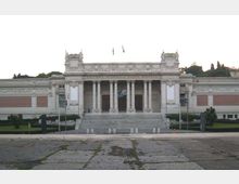 Facciata di un grande edificio neoclassico con alto portico a colonne e ampia scalinata centrale; ai lati si estendono ali simmetriche con decorazioni in pietra e pannelli murari. Davanti c’è una vasta piazza pavimentata, con aiuole e alberi sullo sfondo.