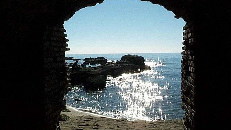 Veduta del mare e di una piccola spiaggia sabbiosa con scogli affioranti, incorniciata dall’apertura ad arco di una struttura in mattoni. L’acqua è illuminata dal riflesso del sole, con l’orizzonte visibile in lontananza.