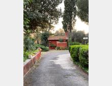 Vialetto asfaltato in un giardino alberato che conduce a un piccolo edificio rosso con tetto in tegole. Ai lati ci sono siepi curate, aiuole e vasi con piante, con alti alberi e lampioni sullo sfondo.