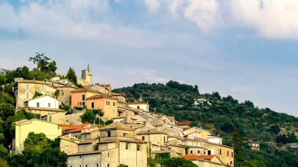 Borgo collinare con case in pietra e facciate color pastello disposte a terrazze, con tetti in coppi e un campanile che spicca tra gli edifici. Intorno si vedono pendii verdi alberati e un ampio cielo parzialmente nuvoloso.