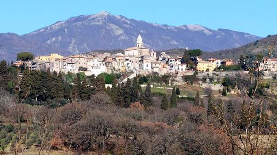Panorama di un borgo collinare con case in pietra e una chiesa con campanile al centro, circondato da alberi e campi. Sullo sfondo si vede una grande montagna sotto un cielo sereno.