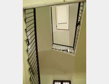 Vano scala interno di un edificio, ripreso dal basso verso l’alto, con rampe sovrapposte e ringhiere metalliche scure che incorniciano un’apertura centrale rettangolare. Pareti chiare e soffitto ad arco, con finestra in fondo che illumina lo spazio.