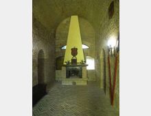 Interno di una sala in pietra con soffitto a volta e pavimento in mattoni disposti a spina di pesce. Al centro c’è un grande camino con cappa alta e chiara, sormontato da uno stemma, con finestre ad arco e torce o supporti lungo le pareti laterali.