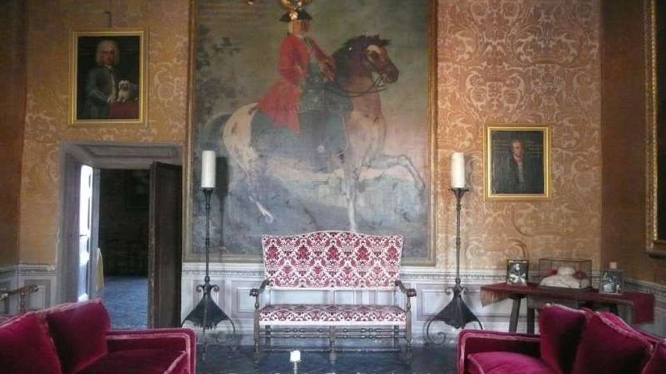 Salone interno in stile storico con pareti rivestite di carta da parati decorata, grande dipinto equestre centrale e ritratti incorniciati ai lati. Al centro una panca imbottita e due alti candelabri, con divani in velluto rosso in primo piano e lampadario sospeso al soffitto.