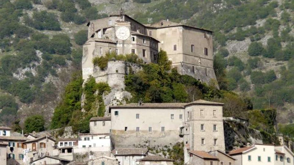 Castello in pietra su uno sperone roccioso sopra un borgo, con un edificio principale dotato di grande orologio sulla facciata e mura di contenimento. Sullo sfondo si vede un pendio montano con vegetazione rada.