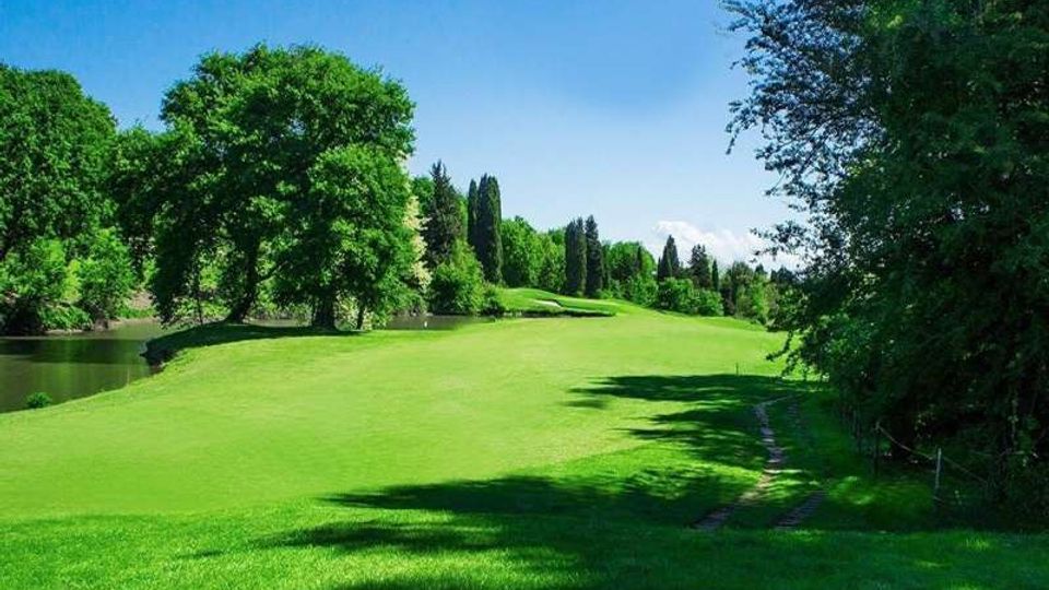 Ampio prato verde ondulato in un parco, costeggiato da un piccolo specchio d’acqua sulla sinistra e circondato da alberi ad alto fusto. In primo piano si vede un sentiero stretto tra l’erba, con cielo sereno sullo sfondo.
