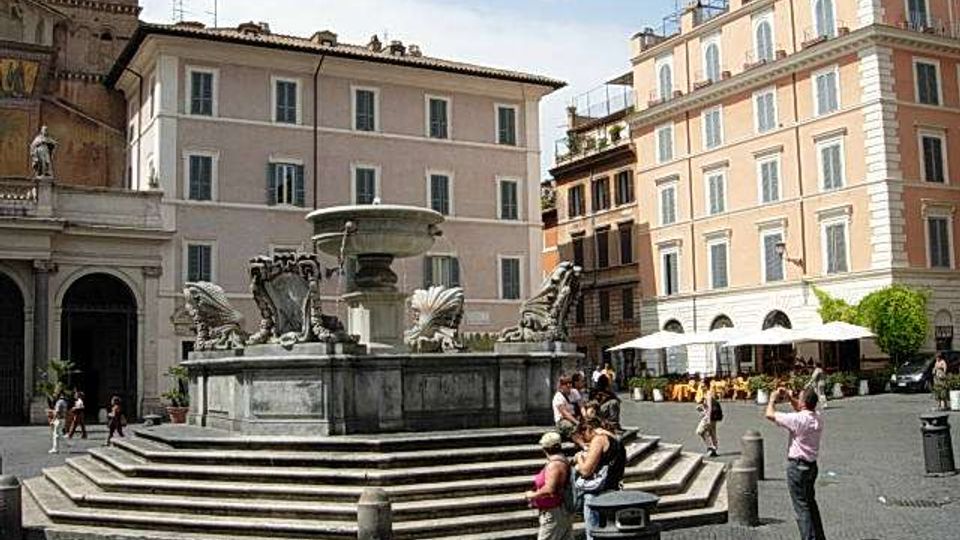Piazza urbana lastricata con una grande fontana monumentale al centro, su basamento a gradoni e sculture decorative. Sullo sfondo si vedono edifici storici a più piani con facciate chiare e finestre regolari, oltre a un angolo di torre con orologio e alcuni ombrelloni di un locale.