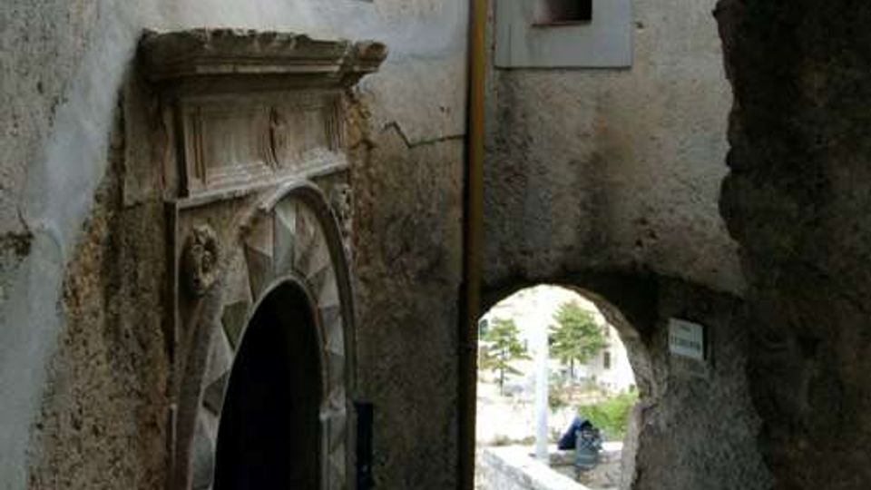 Vicolo stretto in pietra tra edifici in muratura intonacata, con portale ad arco in pietra lavorata e piccole finestre rettangolari. In fondo, un arco passante conduce a una stradina acciottolata e a uno spazio esterno illuminato.