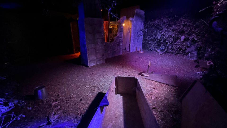 Area esterna notturna in un bosco con illuminazione blu e viola, con un percorso in terra e foglie. Al centro si vedono strutture sceniche in legno e pannelli a forma di rovine, con una luce calda sullo sfondo e un contenitore rettangolare in legno in primo piano.