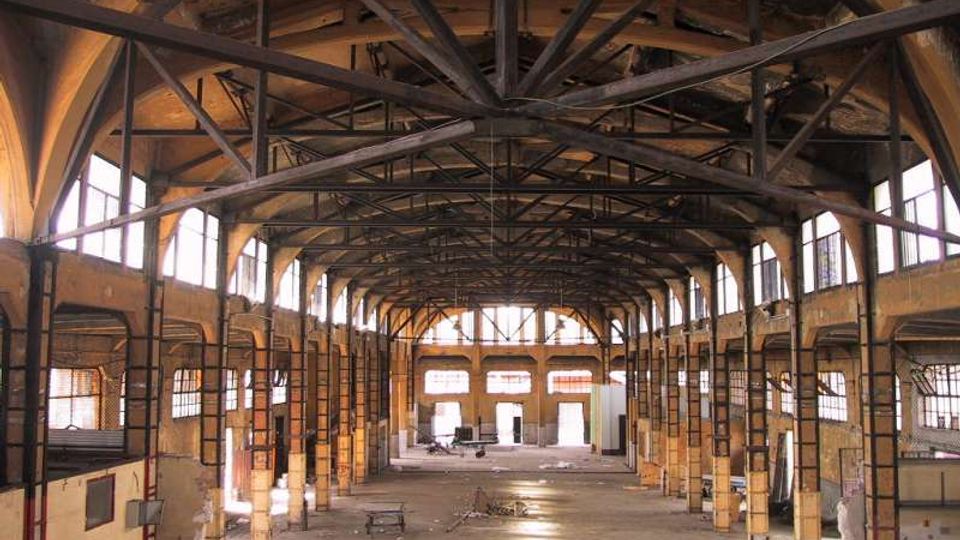 Interno di un grande capannone industriale dismesso, con struttura del tetto in travi metalliche a vista e soffitto alto. Lungo le pareti laterali ci sono file di finestre rettangolari che illuminano un ampio spazio centrale vuoto con pavimento in cemento e pilastri in muratura.