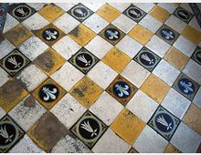 Pavimento in piastrelle a scacchiera con quadrati gialli e bianchi, intervallati da inserti decorativi blu e neri con motivi floreali. Le piastrelle mostrano segni di usura e macchie che evidenziano l’età della superficie.