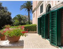 Terrazza esterna di un edificio in stile mediterraneo con facciata chiara e grandi porte-finestre ad arco dotate di persiane verdi aperte. Pavimentazione in piastrelle con fioriere di gerani e giardino con siepi e palme sullo sfondo sotto cielo sereno.