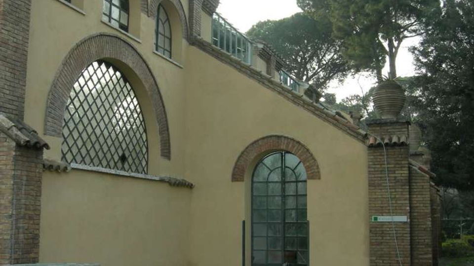 Facciata di edificio con intonaco chiaro e dettagli in mattoni, con grandi finestre ad arco e griglie metalliche. Accanto si vede un pilastro in mattoni con vaso decorativo e alberi alti sullo sfondo.