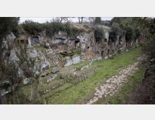 Parete rocciosa con numerose aperture scavate nella pietra, simili a tombe o nicchie, lungo un antico sito rupestre. In primo piano si vede un sentiero lastricato che costeggia una fascia erbosa ai piedi della roccia, con vegetazione bassa in cima.