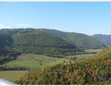 Veduta panoramica di una valle con campi verdi e boschi, circondata da colline ricoperte di vegetazione. Sullo sfondo si estendono rilievi collinari sotto un cielo sereno.