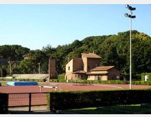 Impianto sportivo all’aperto con pista di atletica rossa e area per il salto in alto, recintato e con un alto palo per l’illuminazione. Sullo sfondo si vedono edifici in mattoni con tetti in coppi e una torre, circondati da alberi e vegetazione.