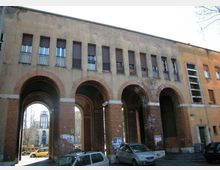 Facciata di un edificio urbano con grandi arcate in mattoni che formano un portico a più passaggi; sopra, una fila di finestre rettangolari con persiane e piccole ringhiere. Sotto le arcate si intravede la strada con auto parcheggiate e, sullo sfondo, una torre.