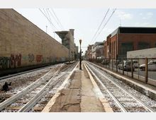 Binari del tram in un corridoio urbano con banchina centrale stretta e lampione, sotto cavi aerei. Ai lati si vedono muri con graffiti, edifici industriali e una recinzione con auto parcheggiate.