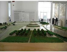 Modello in scala di un parco archeologico con alberi in miniatura, vialetti e una struttura circolare terrazzata al centro. L’allestimento è in una sala museale luminosa con pareti bianche, pannelli informativi e grandi vetrate sul lato destro.