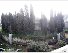Area verde urbana con alti cipressi e altra vegetazione, circondata da un muro in pietra curvo con tratti di recinzione. Sullo sfondo si intravedono edifici residenziali e un piccolo corso d’acqua o fossato ai piedi del muro.