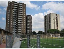 Vista urbana con due alti palazzi residenziali a più piani e balconi sullo sfondo. In primo piano si vede un’area verde recintata con reti metalliche e un passaggio pedonale, sotto un cielo parzialmente nuvoloso.