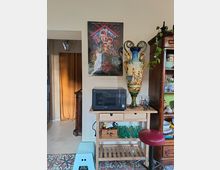Angolo di cucina con carrellino in legno che sostiene un forno a microonde e una grande anfora decorativa, sotto un poster appeso alla parete. A destra c’è una credenza in legno con stoviglie e una pianta ricadente, mentre a sinistra si vede un passaggio con tenda color ocra e lanterna a soffitto.