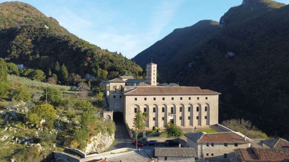 Veduta panoramica di un complesso religioso in pietra, con lunga facciata ad arcate e un campanile quadrato, situato in una valle tra colline boscose. Intorno si vedono strade e piccoli edifici con tetti in coppi, con montagne sullo sfondo.